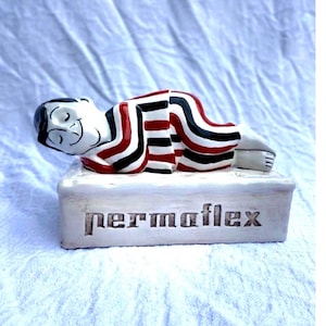Può includere: Statuina in ceramica di una persona sdraiata, dipinta con strisce rosse, nere e bianche. La figura poggia su una base rettangolare bianca con la scritta "permoflex" incisa. La statuina ha un volto sorridente.