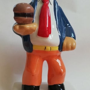 Può includere: Una statuetta in ceramica di un personaggio dei cartoni animati con cappello arancione, giacca blu, camicia bianca, cravatta rossa, cintura gialla e pantaloni arancioni, che tiene in mano un hamburger marrone. La base riporta il testo CHIX, Popeye e Chewing Gum.