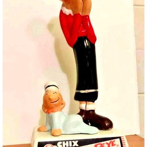Może przedstawiać: Figurka Popeye'a i Oliwki z epoki vintage. Popeye ma na sobie czerwoną kurtkę i czarne spodnie, a Oliwka leży. Na podstawie napis "CHIX art art! POPEYE CARD GUM".