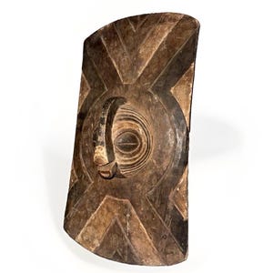 May include: A rectangular wooden shield featuring a dark brown finish with carved geometric patterns. The centre displays a raised, circular relief element with concentric lines and a protruding, curved handle-like structure at the top.