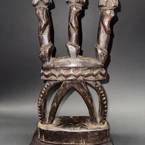 Puede incluir: Un taburete de madera tallada de color marrón oscuro con tres soportes verticales rematados con formas redondeadas. Los soportes están conectados a un asiento circular con un patrón geométrico. El taburete descansa sobre una base rectangular.