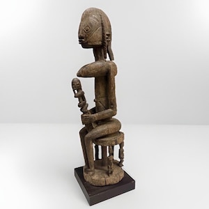 May include: A carved wooden sculpture of a seated figure on a stool, holding a smaller vertical object. The figure has a textured, weathered brown surface with geometric facial features, a prominent crest, and is mounted on a dark square base.