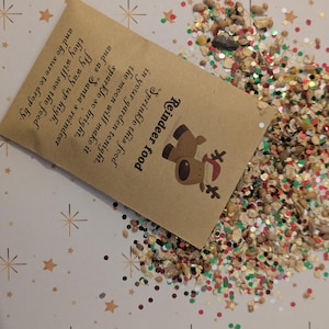 May include: A small brown paper packet of "Reindeer food" with a reindeer illustration, spilling out a mix of grains, seeds, and glitter. The packet's text explains how to sprinkle the food for Santa's reindeer. The background is a light surface with star patterns.