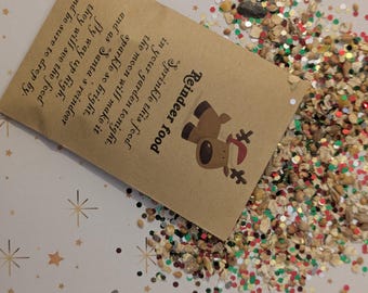 May include: A small brown paper packet of "Reindeer food" with a reindeer illustration, spilling out a mix of grains, seeds, and glitter. The packet's text explains how to sprinkle the food for Santa's reindeer. The background is a light surface with star patterns.