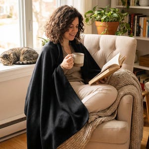 以下が含まれることがあります： ベージュのアームチェアに座り、黒い毛布に包まれ、本を読み、マグカップを持っている人。編み物のスローケットが椅子に掛けられています。窓辺で猫が寝ています。本棚と植物が背景にあります。