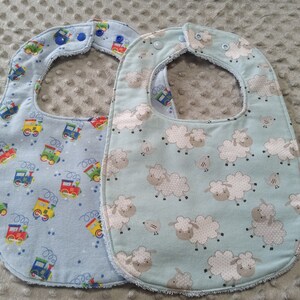 May include: Two baby bibs. One is light blue with colorful train graphics, the other is light teal with white sheep. Both have snap closures at the neck and are made of soft fabric, perfect for mealtime.