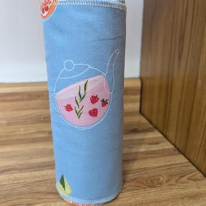 May include: A rolled-up, light blue cloth with a whimsical teapot design. The teapot is pink with red strawberry accents and green leaves. The cloth is approximately 8 inches tall and features a white stitched edge.