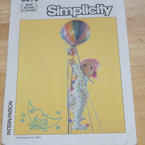 May include: Simplicity sewing pattern featuring a doll holding a rainbow-colored balloon. The doll wears a white onesie with colorful polka dots and pom-poms. The pattern is titled "Moon Babies" with a crescent moon and star graphic.