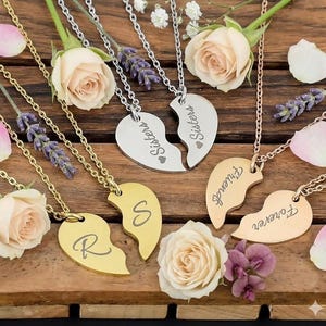 Puede incluir: Collares con colgantes en forma de corazón divididos en dos, con las palabras "Sisters" y "Friends" grabadas. Los collares son dorados y plateados, con cadenas finas. También hay rosas y ramitas de lavanda.