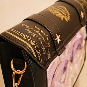 May include: Black handbag with gold accents, shaped like a book. The bag features gold text and star-shaped embellishments. The text reads "Morning Galaxy" and "Close your eyes". The bag has a gold chain and clasp.
