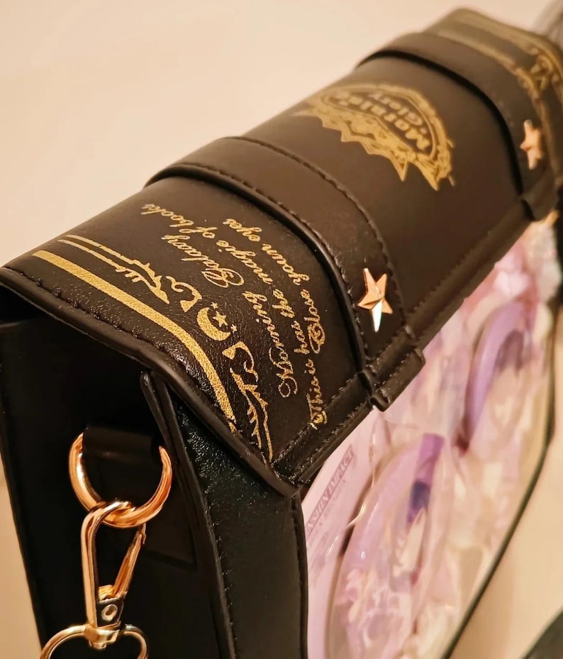 May include: Black handbag with gold accents, shaped like a book. The bag features gold text and star-shaped embellishments. The text reads "Morning Galaxy" and "Close your eyes". The bag has a gold chain and clasp.