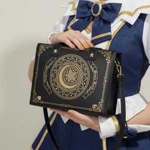 May include: Black handbag with a gold celestial design featuring a crescent moon and star. The bag has a shoulder strap and decorative gold accents. The text on the bag reads "Journey Galaxy" and "Sky of the Moon".