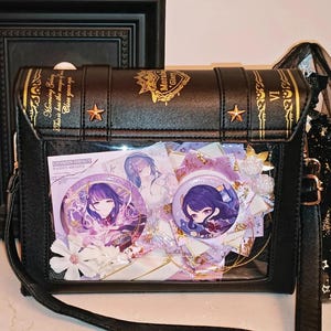 May include: Black crossbody bag with a book-like design, gold accents, and a clear front panel displaying anime-style character pins and decorations. The bag features a long strap and a decorative tassel.
