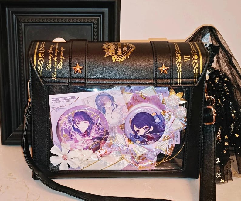 May include: Black crossbody bag with a book-like design, gold accents, and a clear front panel displaying anime-style character pins and decorations. The bag features a long strap and a decorative tassel.