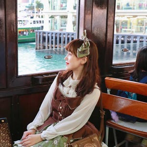 May include: A woman wearing a brown dress with a white blouse and a green skirt, sitting by a window. She has a brown purse with a gold design. The background shows a body of water and buildings.