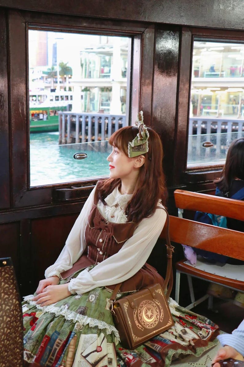 May include: A woman wearing a brown dress with a white blouse and a green skirt, sitting by a window. She has a brown purse with a gold design. The background shows a body of water and buildings.
