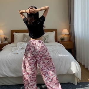 May include: A person wearing a black t-shirt and pink camouflage print pants. The pants have a wide-leg design. The person is standing in front of a bed with white bedding and two bedside tables with lamps.