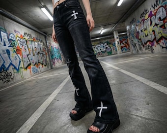 May include: Black denim jeans with white cross embroidery on the front pockets and lower legs. The jeans have a flared leg design and are paired with black platform sandals. The background is a concrete parking garage with graffiti art.