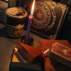 May include: A still-life scene featuring a lit purple candle, a slice of chocolate cake, a Starbucks coffee cup, and a black bag with gold embellishments. A small book and a brown fluffy notebook are also present. The scene is set on a dark surface.