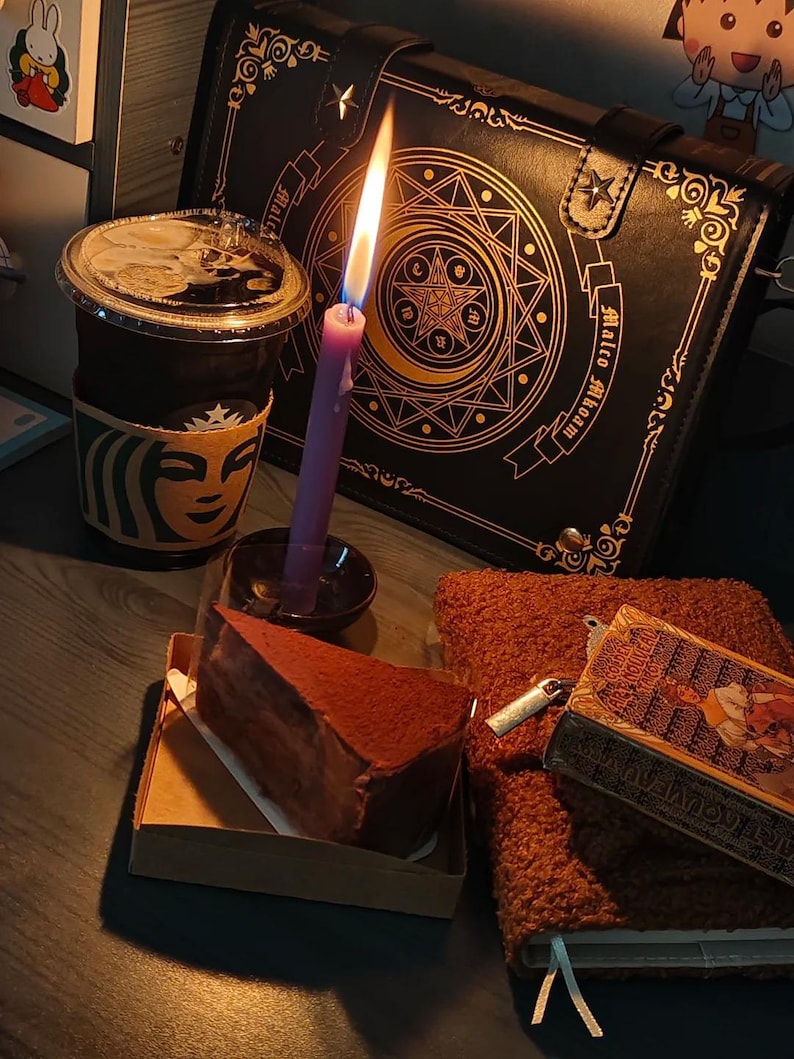 May include: A still-life scene featuring a lit purple candle, a slice of chocolate cake, a Starbucks coffee cup, and a black bag with gold embellishments. A small book and a brown fluffy notebook are also present. The scene is set on a dark surface.