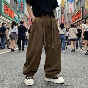 Puede incluir: Pantalones de pana marrones de pernera ancha con cinturón a juego. Los pantalones de cintura alta se llevan con zapatillas blancas. La imagen muestra a una persona en una calle de la ciudad, con gente y tiendas al fondo.