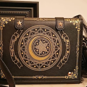May include: Black crossbody bag with a book design. The bag features gold embellishments, including a crescent moon and starburst design, and the words "Magic Book" and "Harmony Galaxy". The bag has a shoulder strap and a decorative tulle accent.