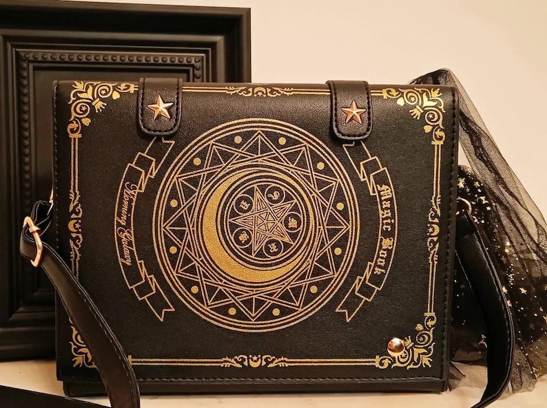 May include: Black crossbody bag with a book design. The bag features gold embellishments, including a crescent moon and starburst design, and the words "Magic Book" and "Harmony Galaxy". The bag has a shoulder strap and a decorative tulle accent.