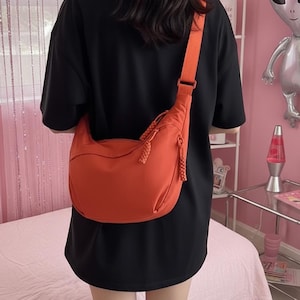 May include: An orange shoulder bag with a wide strap and a curved design. The bag has a braided detail on the zipper pulls and is worn over the shoulder. The person in the image is wearing a black t-shirt.