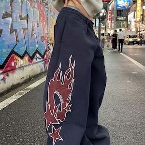 May include: Navy blue sweatpants with a flame and star graphic in burgundy and white. The text "COBB&MASS" is within the flame design. The pants have a relaxed fit and a drawstring waist.