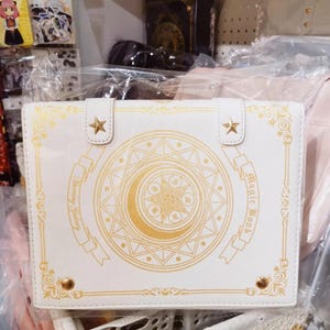 May include: White faux leather book-style clutch with gold accents. The cover features a gold crescent moon, star, and geometric design, with the words "Magic Book" and "Blooming Galaxy". Two gold star-shaped clasps secure the top.