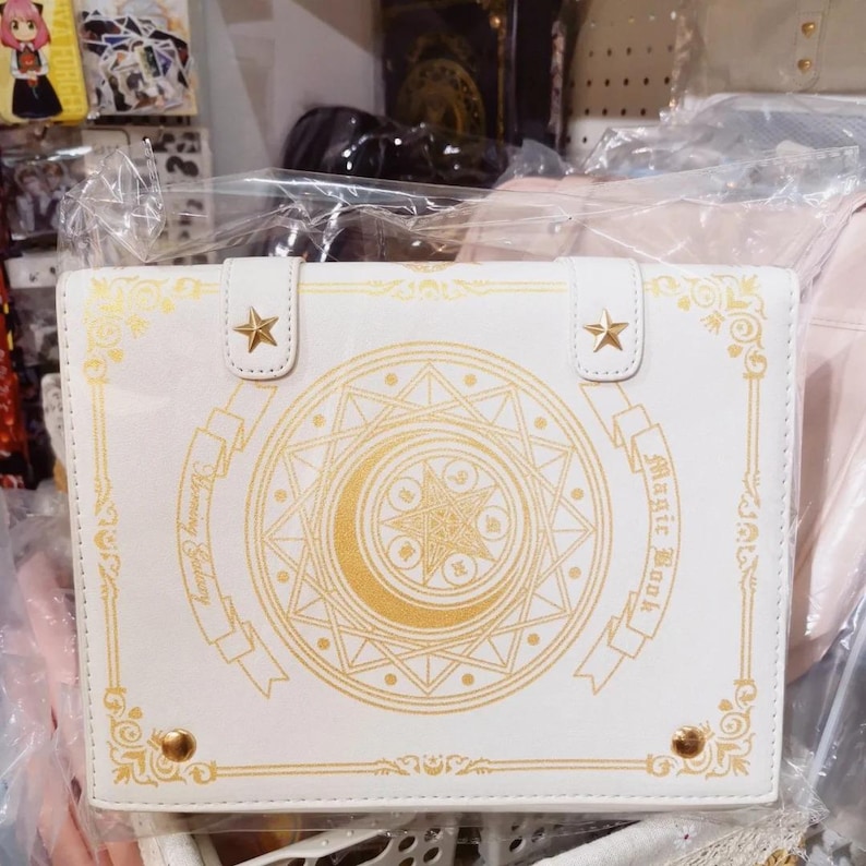 May include: White faux leather book-style clutch with gold accents. The cover features a gold crescent moon, star, and geometric design, with the words "Magic Book" and "Blooming Galaxy". Two gold star-shaped clasps secure the top.