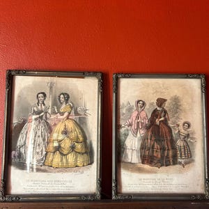 Peut inclure: Deux gravures de mode anciennes encadrées. Les gravures présentent des illustrations détaillées de femmes en robes et bonnets élaborés. Les cadres sont sombres avec des détails ornés. Le fond est un mur rouge uni.