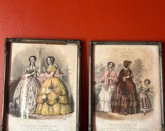 Könnte beinhalten: Zwei gerahmte antike Mode-Drucke. Die Drucke zeigen detaillierte Illustrationen von Frauen in aufwendigen Kleidern und Hauben. Die Rahmen sind dunkel mit aufwendigen Details. Der Hintergrund ist eine einfarbige rote Wand.
