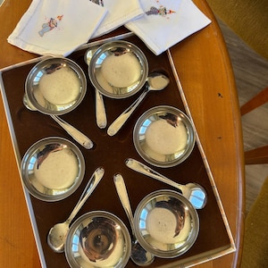 Peut inclure: Un ensemble de petits bols et cuillères argentés, disposés dans une boîte marron. Les bols sont ronds avec une petite poignée. Des serviettes en lin blanc avec des figures brodées sont également incluses.