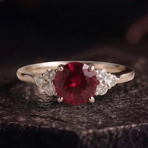 Könnte beinhalten: Ein goldener Ring mit einem großen, runden, tiefroten Edelstein in der Mitte, flankiert von drei kleineren, klaren, rund geschliffenen Steinen auf jeder Seite. Die Ringschiene ist glatt und poliert und zeigt eine klassische Krappenfassung.