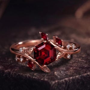 Könnte beinhalten: Ein Ring aus Roségold mit einem zentralen hexagonalen roten Edelstein, umgeben von kleineren roten Steinen im Marquise-Schliff und winzigen klaren runden Akzentsteinen. Das Band ist mit zarten Metallblattmotiven verziert.