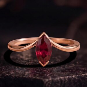 Könnte beinhalten: Ein Ring aus Roségold mit einem tiefroten Edelstein im Marquise-Schliff, der in eine V-förmige Ringschiene eingefasst ist. Das Metall hat eine glatte, polierte Oberfläche und der Stein ist vertikal in der Mitte platziert und eingefasst.