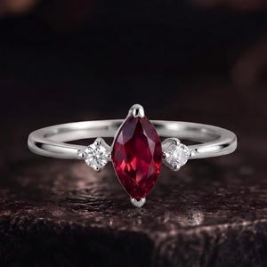 Può includere: Un anello color argento con una gemma centrale rosso scuro taglio marquise, affiancata da due piccole pietre bianche rotonde e trasparenti. La fascia è liscia e lucida, caratterizzata da un classico design a tre pietre.
