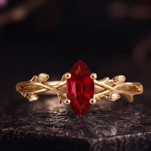 Könnte beinhalten: Ein goldener Ring mit einem zentralen, marquise-geschliffenen roten Edelstein. Die Ringschiene ist mit einem strukturierten, astähnlichen Motiv gestaltet, das kleine, geformte Blätter und Knospen umfasst, die von der Fassung ausgehen.
