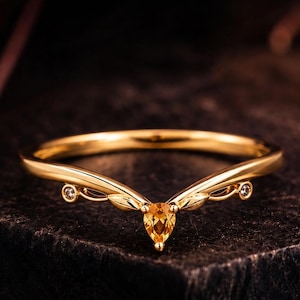 Puede incluir: Un anillo dorado en forma de V con una piedra preciosa amarilla central en talla de pera. La banda presenta un delicado diseño inspirado en hojas con dos pequeñas piedras redondas transparentes a cada lado, creando una silueta simétrica.