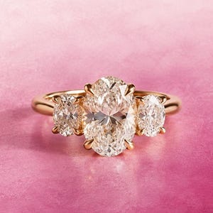 Puede incluir: Un anillo de oro con un gran diamante central de corte ovalado flanqueado por dos diamantes más pequeños de corte ovalado. Las tres piedras están montadas en una banda de oro pulido, creando un diseño clásico de tres piedras facetadas.
