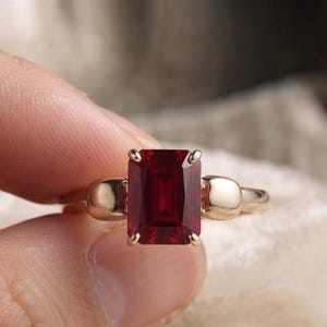Pu&ograve; includere: Un anello con una grande pietra preziosa rettangolare rosso scuro incastonata in una fascia di metallo lucido color oro. L'incastonatura include due dettagli metallici lisci e arrotondati su entrambi i lati della pietra centrale, fissata da quattro griffe.