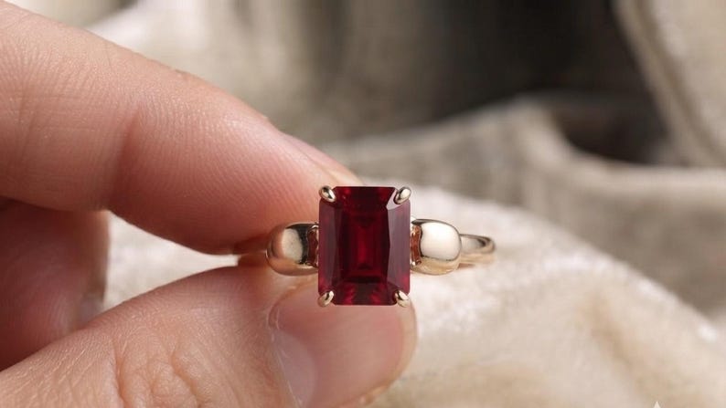 Pu&ograve; includere: Un anello con una grande pietra preziosa rettangolare rosso scuro incastonata in una fascia di metallo lucido color oro. L'incastonatura include due dettagli metallici lisci e arrotondati su entrambi i lati della pietra centrale, fissata da quattro griffe.