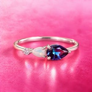 Könnte beinhalten: Ein silberfarbener Ring mit einem kleinen runden klaren Stein, einem birnenförmigen schillernden Opal und einem größeren birnenförmigen, dunkelblau und violett changierenden Stein, gefasst in eine zarte Metallschiene mit poliertem Finish.