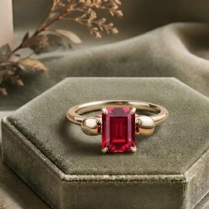 Pu&ograve; includere: Un anello in oro con una pietra preziosa rosso intenso dal taglio rettangolare a smeraldo al centro. La fascia presenta due piccole sfere d'oro lisce su entrambi i lati della pietra centrale, creando un design equilibrato e simmetrico.