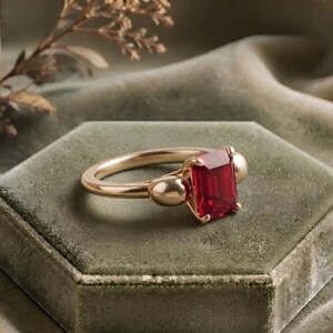 Pu&ograve; includere: Un anello in oro con una pietra preziosa rossa di taglio smeraldo rettangolare al centro, affiancata da due dettagli dorati lisci e arrotondati su entrambi i lati. La fascia &egrave; in oro lucido, conferendo al gioiello un aspetto raffinato.