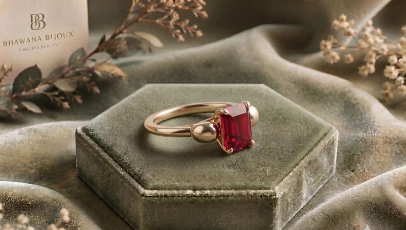 Pu&ograve; includere: Un anello in oro con una pietra preziosa rossa di taglio smeraldo rettangolare al centro, affiancata da due dettagli dorati lisci e arrotondati su entrambi i lati. La fascia &egrave; in oro lucido, conferendo al gioiello un aspetto raffinato.