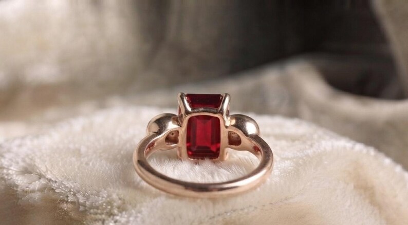 Pu&ograve; includere: Un anello in oro rosa con una pietra preziosa rosso intenso dal taglio rettangolare a smeraldo come pietra centrale. Il gambo dell'anello &egrave; liscio e lucido, con due piccole pietre rotonde incastonate ai lati della pietra centrale.