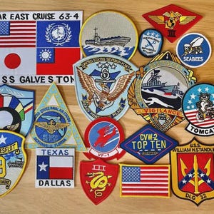 May include: A selection of embroidered patches with military and naval designs. The patches display flags, eagles, ships, and text like "FAR EAST CRUISE 63-4", "USS GALVESTON", and "TOMCAT". The patches use various colours, including red, blue, yellow, and white.