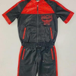 May include: Black and red track suit with the MCM logo. The jacket has a red upper section and sleeves, with black on the lower half. The shorts are black with red side stripes. The suit is made of a leather-like material.